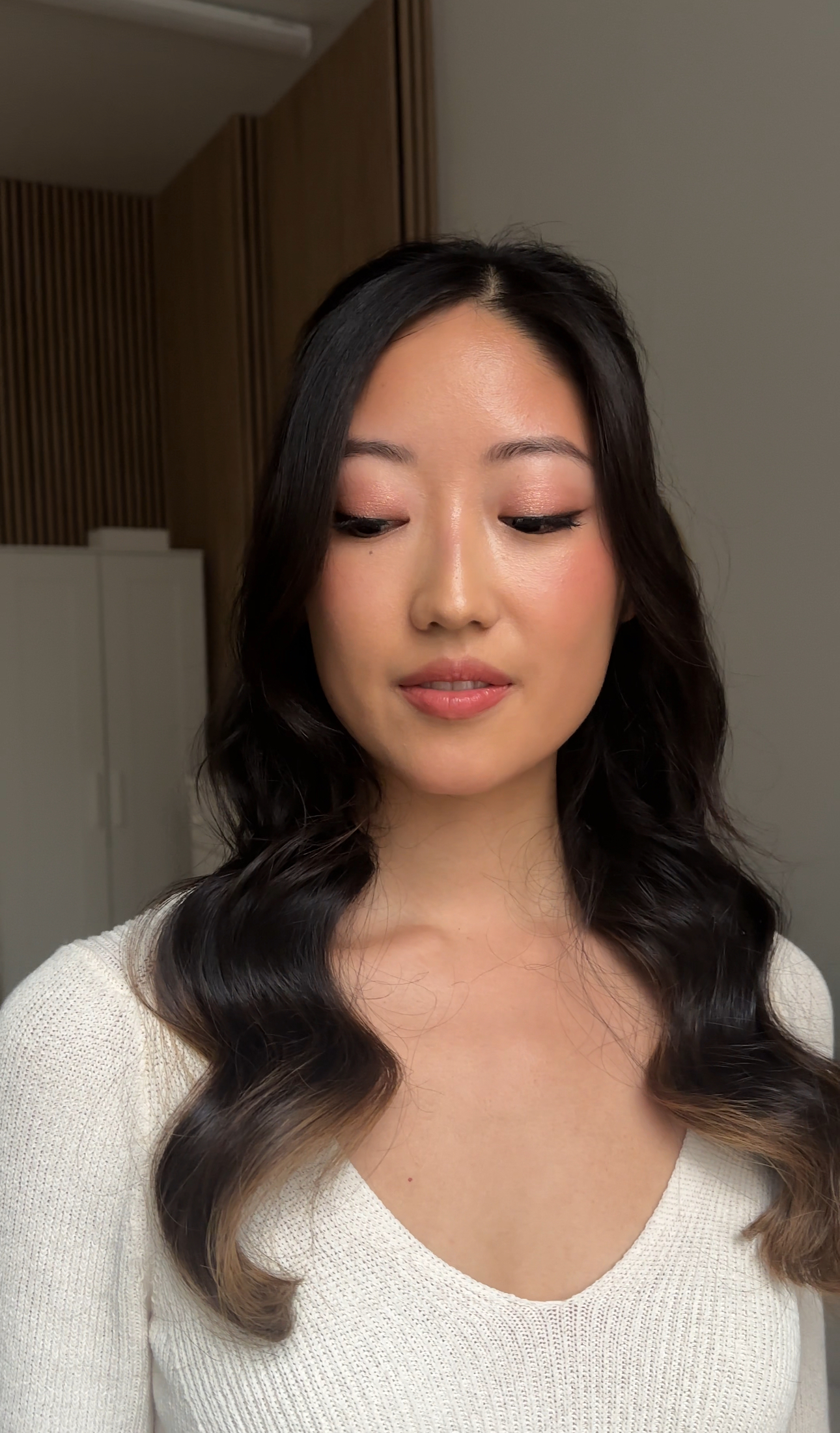 Profile portrait: soft blush, subtle liner, dark waves; cream knit top.