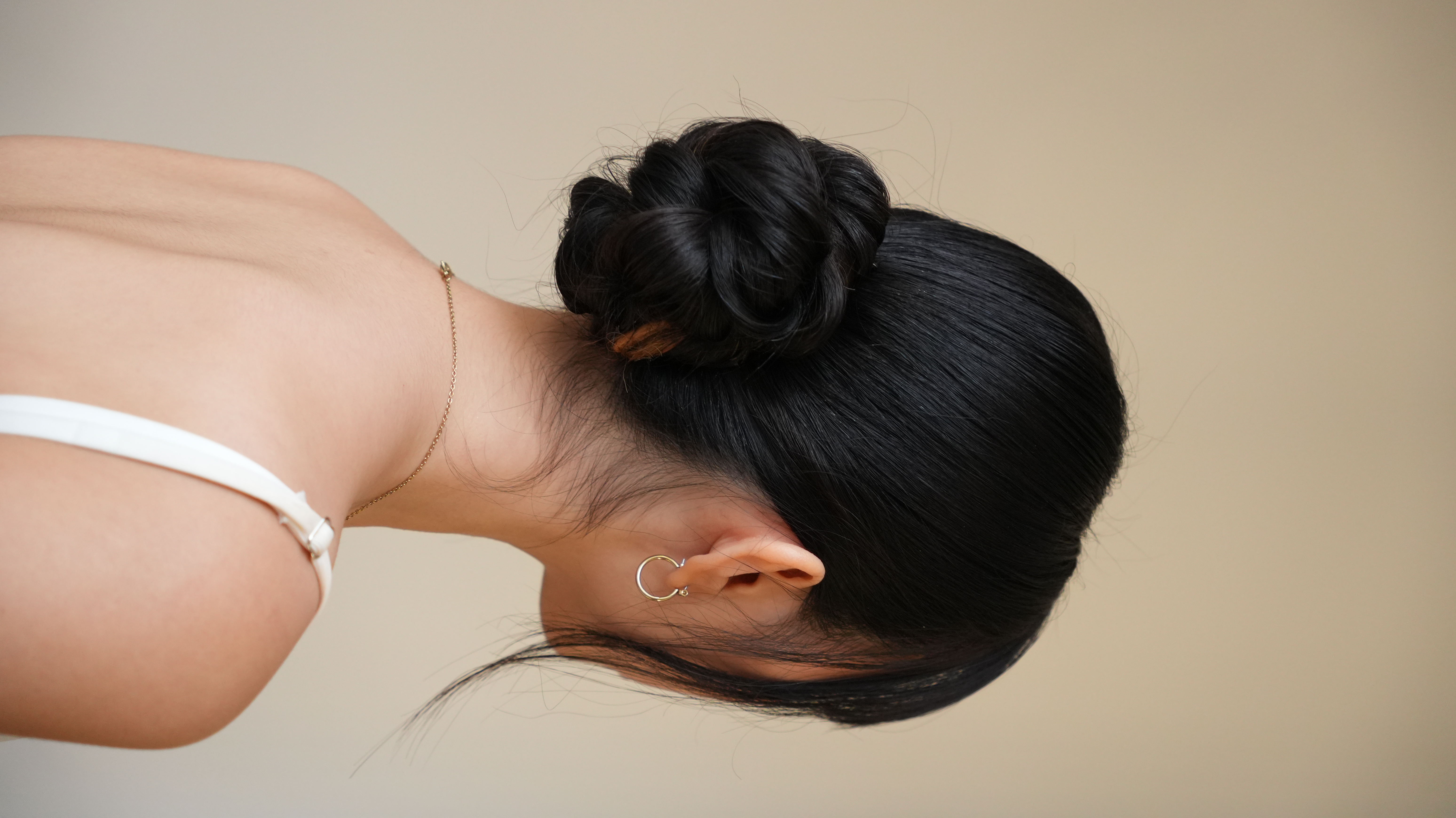Side view: black hair in a low bun, white strap, minimal backdrop.